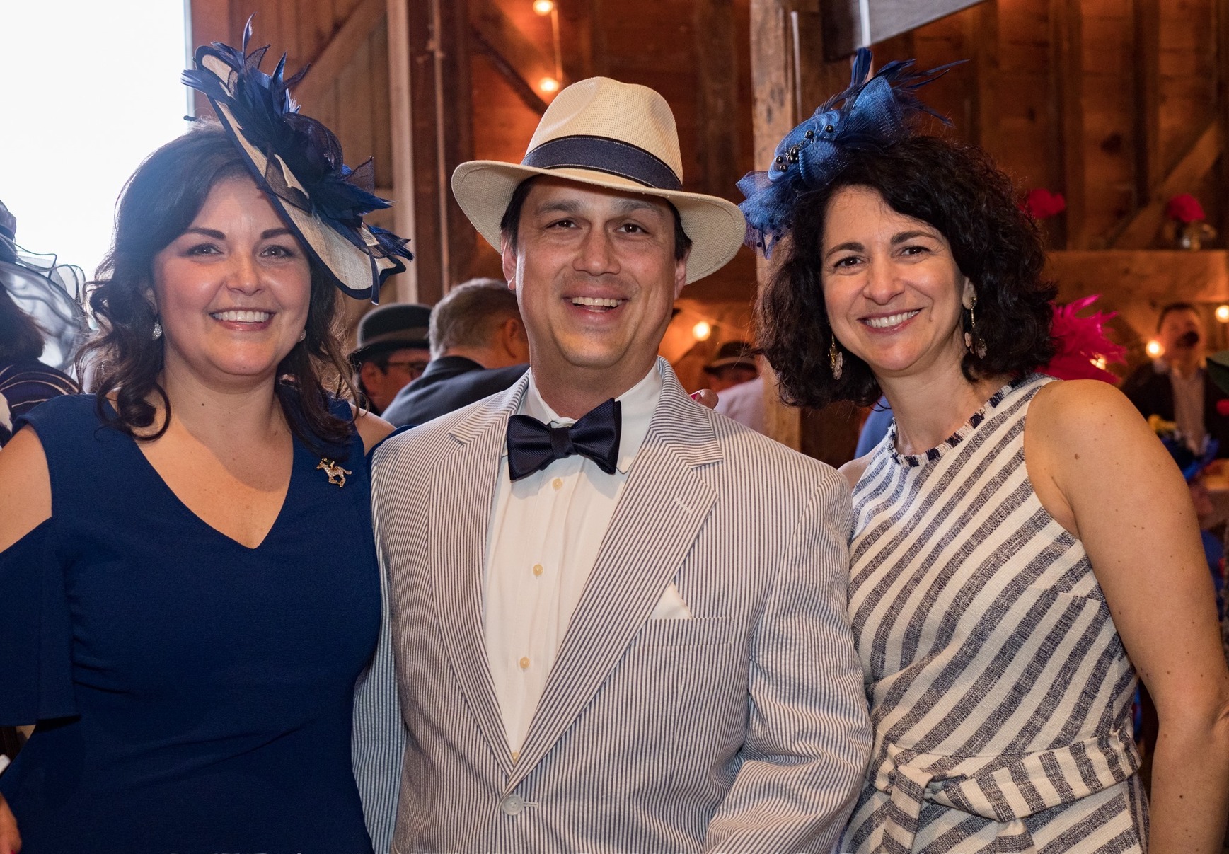 This photo shows a happy group of event attendees. There are two women and one man. They are enjoying themselves at the event. Both women wear big beautiful Kentucky Derby hats.