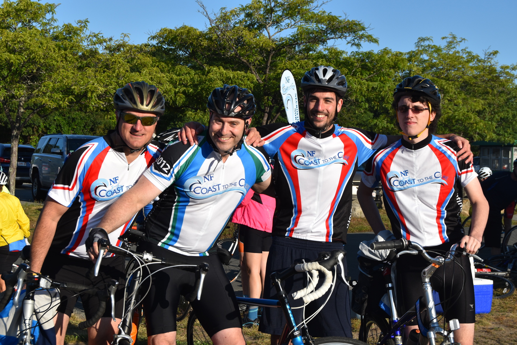 This photograph shows four men posing with Coast to the Cure cycling jerseys on and with their bikes.