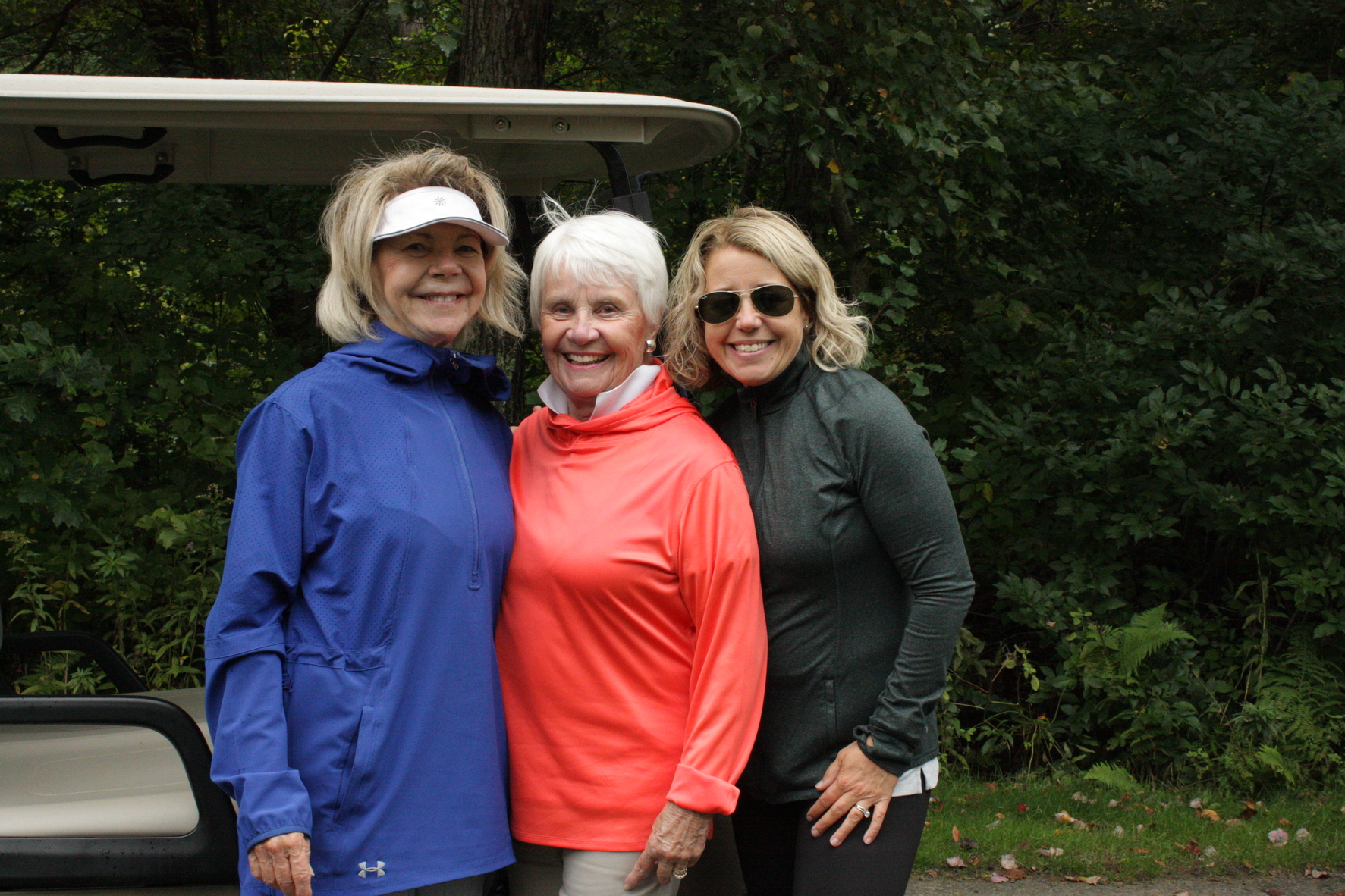 three women in golf-wear posing together in front of a golf cart and wooded land