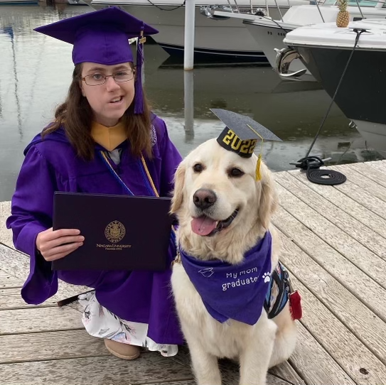 image of girl in purple graduation gown with her service dog in a graduation cap