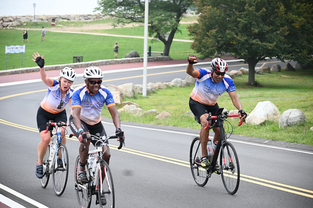 three smiling cyclists biking up a hill, two are waving hello to the camera