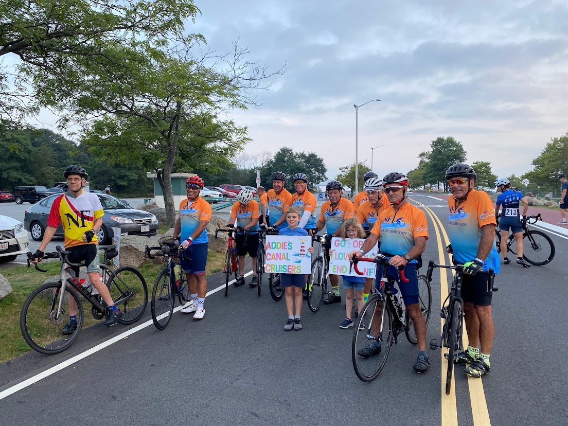 group of cyclists at a start line with two children sitting on handlebars and holding signs that say "Addie's Canal Cruisers"