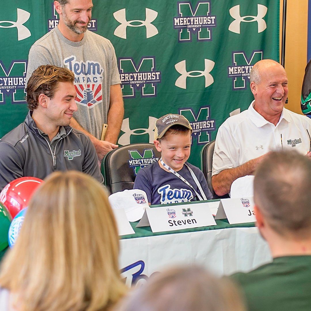 young boy in Team IMPACT gear smiling and signing with Mercyhurst Men's Hockey team, surrounded by coaches and teammates
