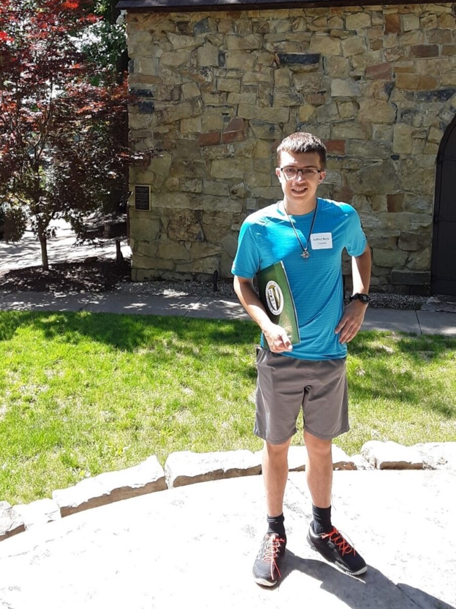 boy in blue shirt and grey shorts stands in front of stone building and grass with folder in hand