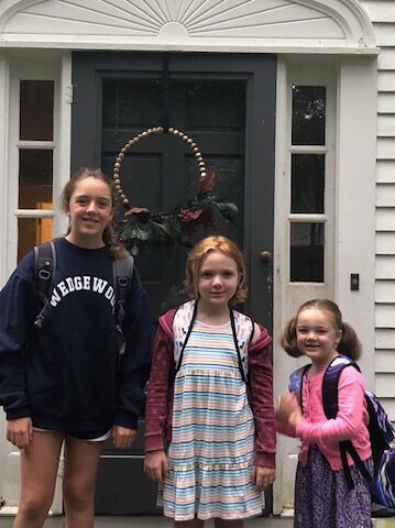 three young girls posing with backpacks on their front stoop on the first day of school; Fiona is in the middle wearing a striped dress