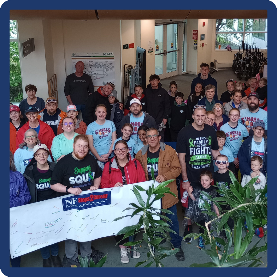 Large group of families, children, and supporters gathered indoors at an NF Northeast Steps2Cure NF event in Lebanon, New Hampshire, holding a signed event banner and wearing neurofibromatosis awareness shirts.